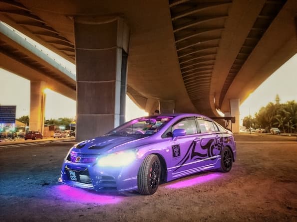 A lowered purple car with neon underglow parked under a highway overpass